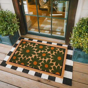 May include: Rectangular doormat with a retro floral design. The mat features large green flowers with orange centers on a tan background. It is framed by a brown border and sits atop a black and white striped rug.