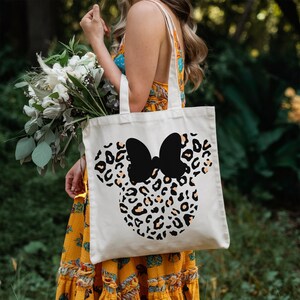 Puede incluir: Un bolso tote de color crema con un dise&ntilde;o de Minnie Mouse. El dise&ntilde;o incluye un lazo negro y un estampado de leopardo en tonos negros, beige y naranja. El bolso lo lleva una persona con un vestido amarillo, con un ramo de flores.