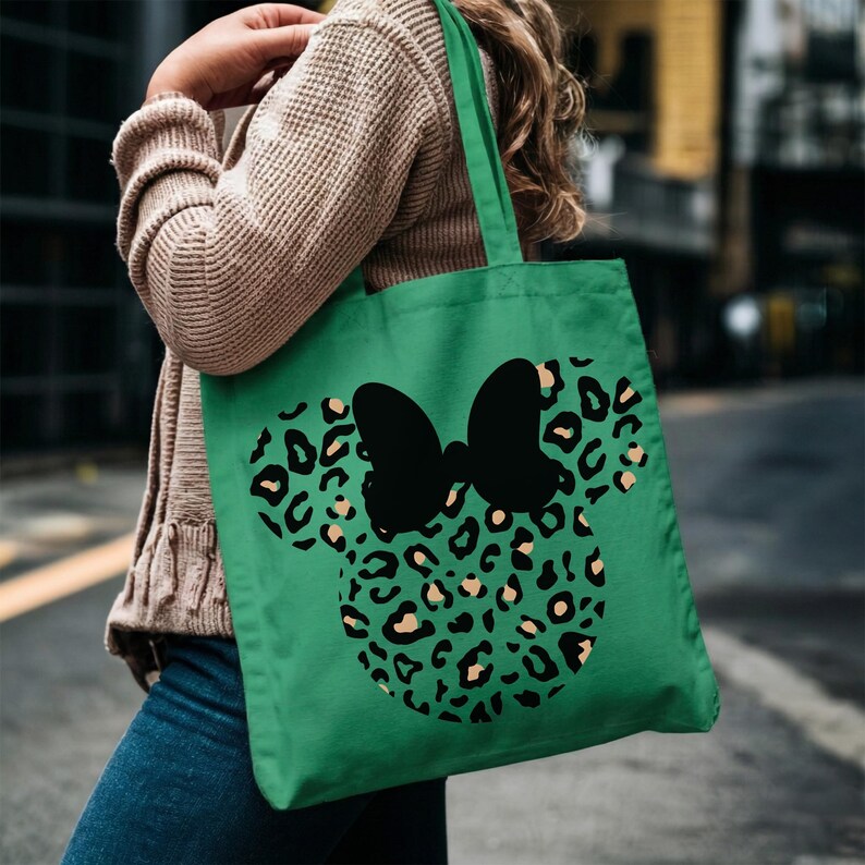 Puede incluir: Bolso tote verde con un lazo negro y un dise&ntilde;o de cabeza de rat&oacute;n con estampado de leopardo. La bolsa est&aacute; hecha de tela duradera y tiene un asa. La bolsa la lleva una persona.