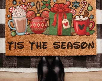 Könnte beinhalten: Eine rechteckige Fußmatte mit festlichem Weihnachtsdesign. Die Matte zeigt eine Tasse heiße Schokolade, Ornamente, ein verpacktes Geschenk und den Text "TIS THE SEASON". Das Design enthält Mickey Mouse-Icons und festliche Farben.