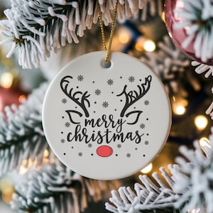 Puede incluir: Adorno navideño redondo de cerámica blanca con una cuerda dorada para colgar. Presenta cuernos de reno negros, copos de nieve y las palabras "merry christmas". Un círculo rojo representa la nariz del reno.