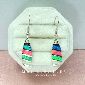 May include: Silver earrings with colorful, layered, rectangular pendants. The pendants feature black, blue, pink, green, and white sections. The earrings are displayed on a white, velvet-like surface. The text "MALIK JEWELER Handcrafted Jeweller" is visible.