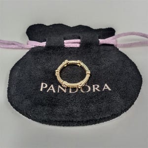 Może przedstawiać: Złoty pierścionek z małymi, przezroczystymi kamieniami leży na czarnym woreczku z białym napisem "PANDORA". Pierścionek ma powtarzający się wz&oacute;r i jest centralnym punktem obrazu.