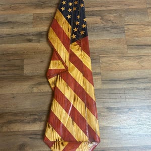 May include: A wooden American flag with red and yellow stripes and a blue field with yellow stars. The flag is carved and has a rustic, handcrafted appearance. The flag is displayed on a wooden surface.
