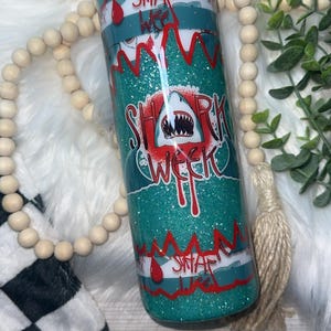 May include: Glittery teal tumbler with a "Shark Week" design. The design features a shark with an open mouth, red accents, and the words "Shark Week" in a dripping font. The tumbler also has the text "Snap" and "Week".