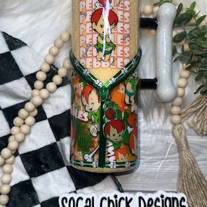 May include: A glittery tumbler featuring Pebbles from The Flintstones. The design includes Pebbles in a green outfit, the word "Pebbles" repeated, and a green handle. The tumbler has a bone-shaped handle and the text "SOCAL Chick Designs".