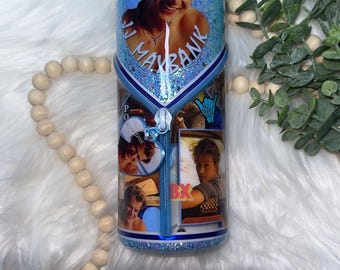 Puede incluir: Un vaso cilíndrico con acabado de purpurina azul que presenta un collage de fotografías de un joven, el texto JJ MAYBANK y un gráfico central de cremallera vertical. El diseño incluye varias imágenes de la misma persona y un icono de mano shaka azul.