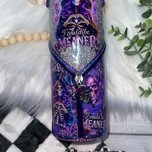 May include: A purple and silver tumbler with a skeleton design. The tumbler features the text "I could be MEANER" and a zipper design with a skull charm. The bottom of the tumbler has the text "socalchickdesigns".
