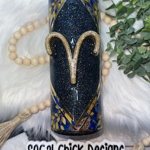 May include: A glittery tumbler with an Aries zodiac symbol. The tumbler features a dark blue glitter background with gold accents and a gold Aries symbol. The design includes floral and geometric patterns. The text "SOCAL Chick Designs" is visible.
