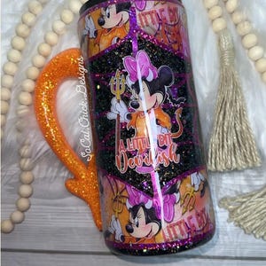 May include: A glittery tumbler with an orange handle. The tumbler features a cartoon Minnie Mouse dressed as a devil, holding a trident. The text "A Little Bit Devilish" is also visible on the tumbler.