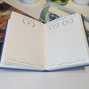 May include: An open blue planner with cream-coloured pages featuring lined sections for daily entries. The left page shows Vendredi 8 Janvier and the right page shows Samedi 9 Janvier and Dimanche 10 Janvier. The text includes Semaine 1 and 2027.