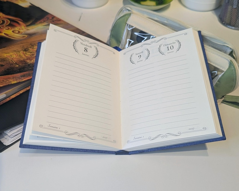 May include: An open blue planner with cream-coloured pages featuring lined sections for daily entries. The left page shows Vendredi 8 Janvier and the right page shows Samedi 9 Janvier and Dimanche 10 Janvier. The text includes Semaine 1 and 2027.