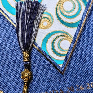 May include: A blue fabric-covered agenda with the text AGENDA 2026 visible. A decorative tassel in navy and white threads hangs from a gold-toned metal charm featuring a flower and a sunburst design, resting against a white card with gold and teal circles.