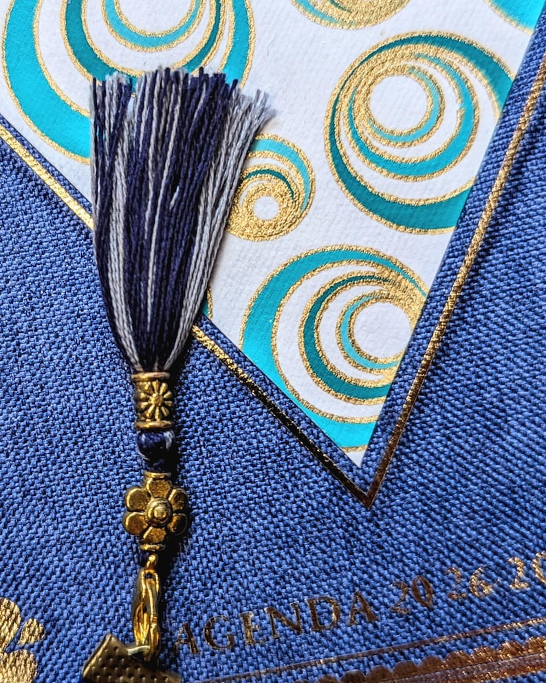 May include: A blue fabric-covered agenda with the text AGENDA 2026 visible. A decorative tassel in navy and white threads hangs from a gold-toned metal charm featuring a flower and a sunburst design, resting against a white card with gold and teal circles.