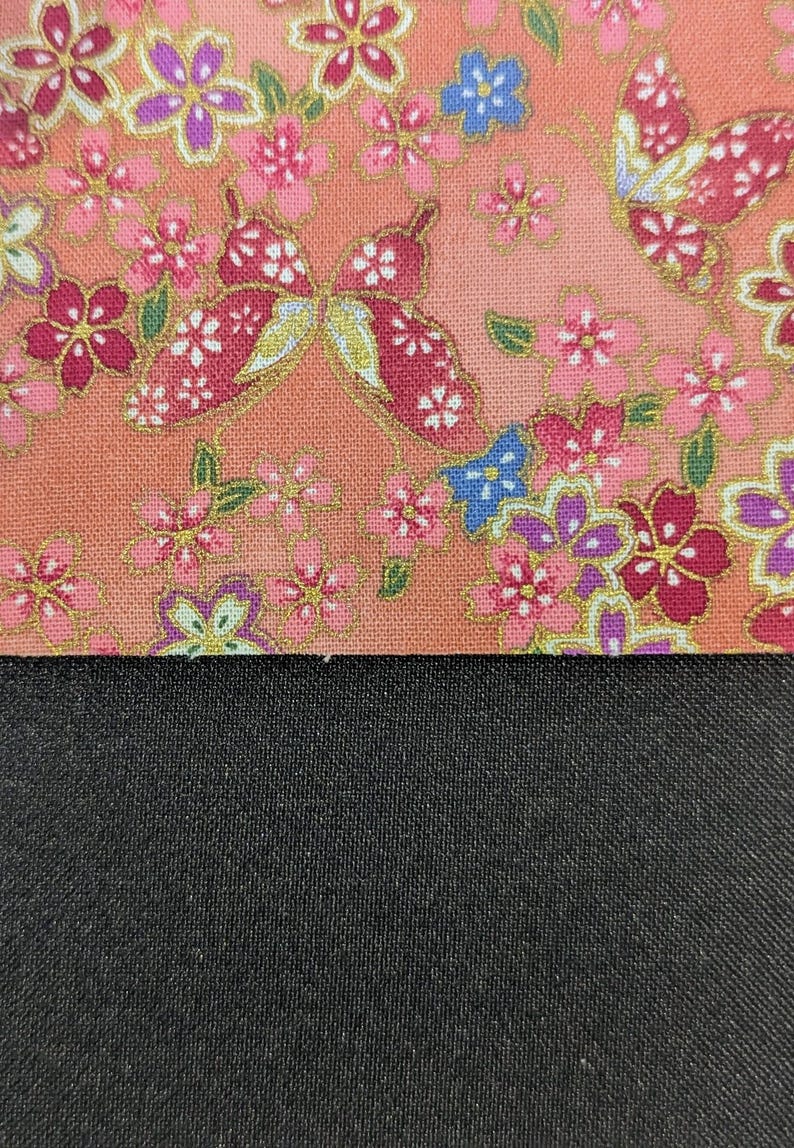 May include: A fabric swatch featuring a peach-coloured background with a repeating pattern of pink, purple, and blue flowers, green leaves, and red butterflies with gold accents, positioned above a solid, textured black fabric section.