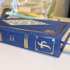 May include: A blue fabric-covered planner with gold foil accents on the spine and cover. The spine displays the years 2026 and 2027 in gold text. The cover features a geometric pattern of teal and white circles and a blue ribbon bookmark.