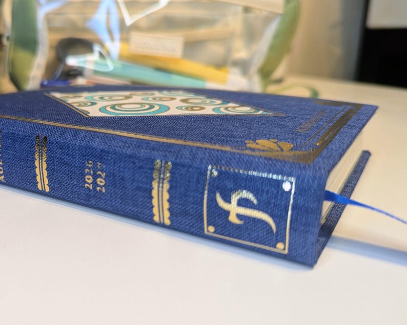May include: A blue fabric-covered planner with gold foil accents on the spine and cover. The spine displays the years 2026 and 2027 in gold text. The cover features a geometric pattern of teal and white circles and a blue ribbon bookmark.