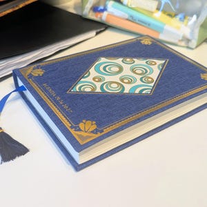 May include: A blue fabric-covered agenda with gold foil corner accents and a central diamond-shaped panel featuring a teal and gold circular pattern. The cover text reads "AGENDA 2026 2027". A blue ribbon bookmark with a dark blue tassel is attached.
