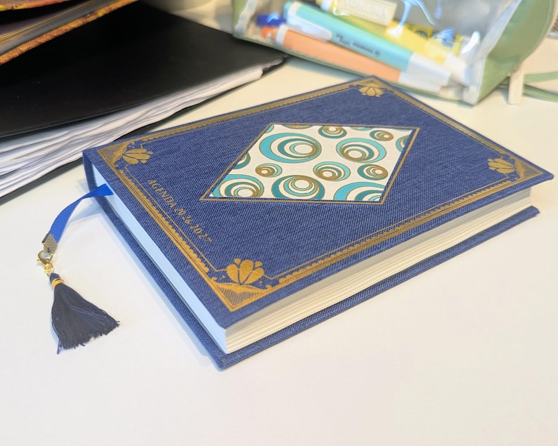 May include: A blue fabric-covered agenda with gold foil corner accents and a central diamond-shaped panel featuring a teal and gold circular pattern. The cover text reads "AGENDA 2026 2027". A blue ribbon bookmark with a dark blue tassel is attached.