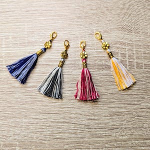 May include: Four decorative tassels with gold-toned lobster claw clasps and flower-shaped metal beads. The tassels feature threads in blue, black and white, magenta and white, and yellow and white, each finished with a small gold-toned metal coil.