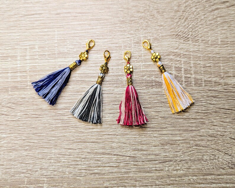 May include: Four decorative tassels with gold-toned lobster claw clasps and flower-shaped metal beads. The tassels feature threads in blue, black and white, magenta and white, and yellow and white, each finished with a small gold-toned metal coil.