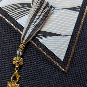 May include: A decorative tassel featuring black and white threads, accented with a gold-toned metal bead and a small flower-shaped charm. The tassel is attached to a gold-toned lobster clasp, which connects to a white fabric element with a gold-toned cap.