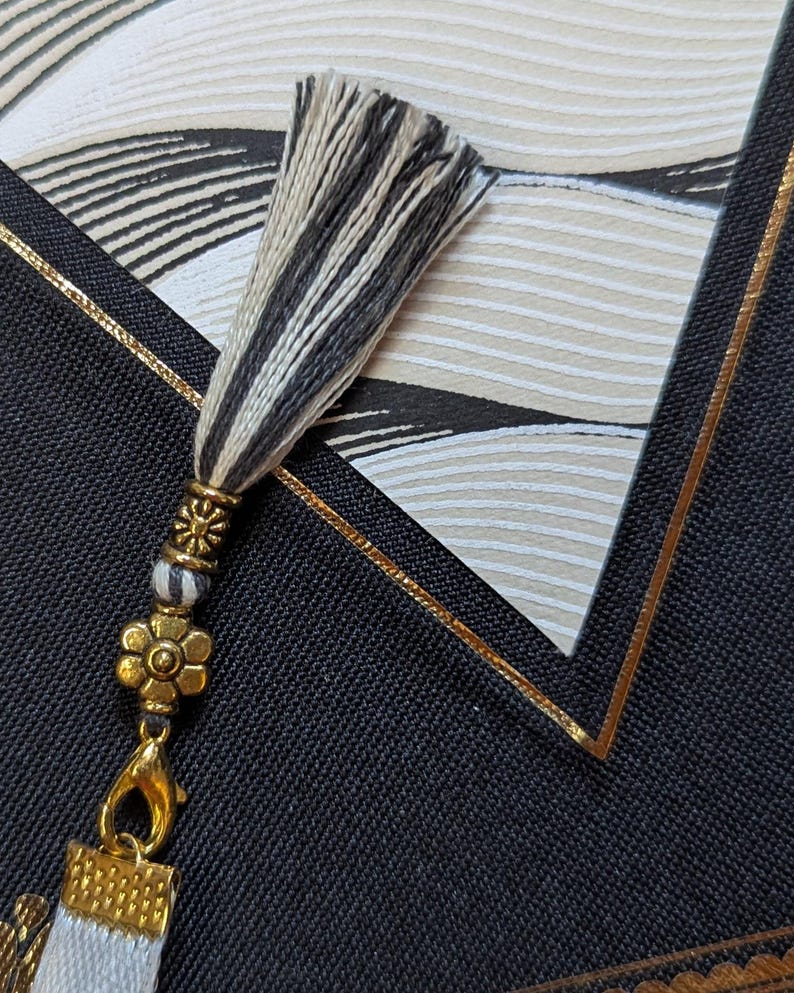 May include: A decorative tassel featuring black and white threads, accented with a gold-toned metal bead and a small flower-shaped charm. The tassel is attached to a gold-toned lobster clasp, which connects to a white fabric element with a gold-toned cap.