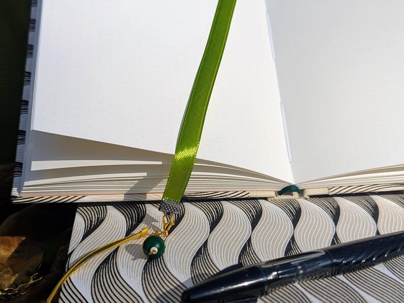K&ouml;nnte beinhalten: Ein offenes Notizbuch mit wei&szlig;en, gepunkteten Seiten verf&uuml;gt &uuml;ber ein gr&uuml;nes Satinband als Lesezeichen, das an einer silbernen Metallklemme befestigt ist. Ein goldfarbener Karabinerverschluss verbindet das Band mit einer dunkelgr&uuml;nen Kugelperle.