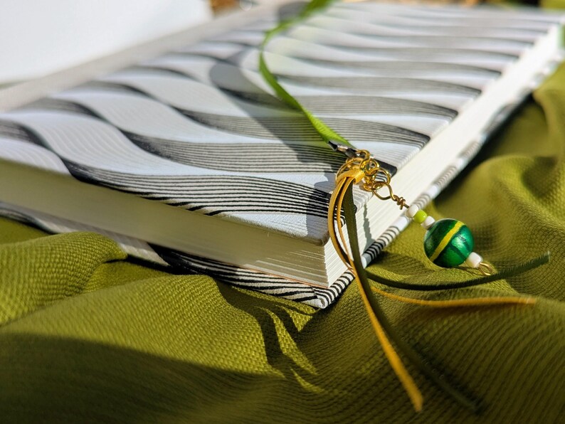 K&ouml;nnte beinhalten: Ein Buch mit wei&szlig;em Einband, schwarzen Wellenlinien und einem gr&uuml;nen Lesezeichenband an einem goldenen Metallring. Das Lesezeichen ist mit einer runden gr&uuml;nen Perle mit gelbem Streifen, kleinen wei&szlig;en Perlen und gelben Lederb&auml;ndern verziert.