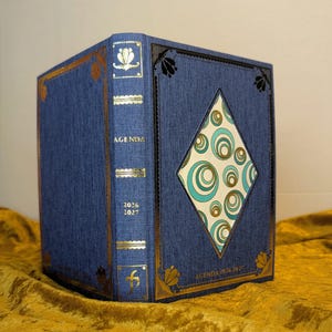 May include: A blue fabric-covered agenda with gold foil accents and a diamond-shaped cutout on the front cover. The cover features a repeating pattern of teal and gold circles. The spine is labelled with the text AGENDA 2026 2027 in gold lettering.