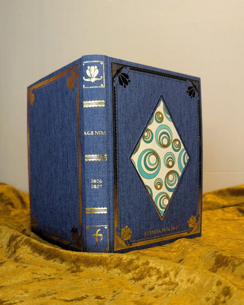 May include: A blue fabric-covered agenda with gold foil accents and a diamond-shaped cutout on the front cover. The cover features a repeating pattern of teal and gold circles. The spine is labelled with the text AGENDA 2026 2027 in gold lettering.