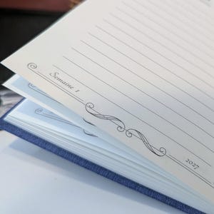 May include: A blue fabric-covered planner with cream-coloured pages. The open page features horizontal lines for writing, decorative black scrollwork borders, and the printed text "Semaine 1" on the left and "2027" on the right side of the page.