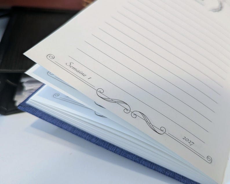 May include: A blue fabric-covered planner with cream-coloured pages. The open page features horizontal lines for writing, decorative black scrollwork borders, and the printed text "Semaine 1" on the left and "2027" on the right side of the page.