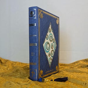 May include: A blue fabric-covered planner with a diamond-shaped centre panel featuring teal and gold concentric circles. The spine and cover are accented with gold foil detailing, including the text "AGENDA 2024-2025". A dark blue tassel is attached.