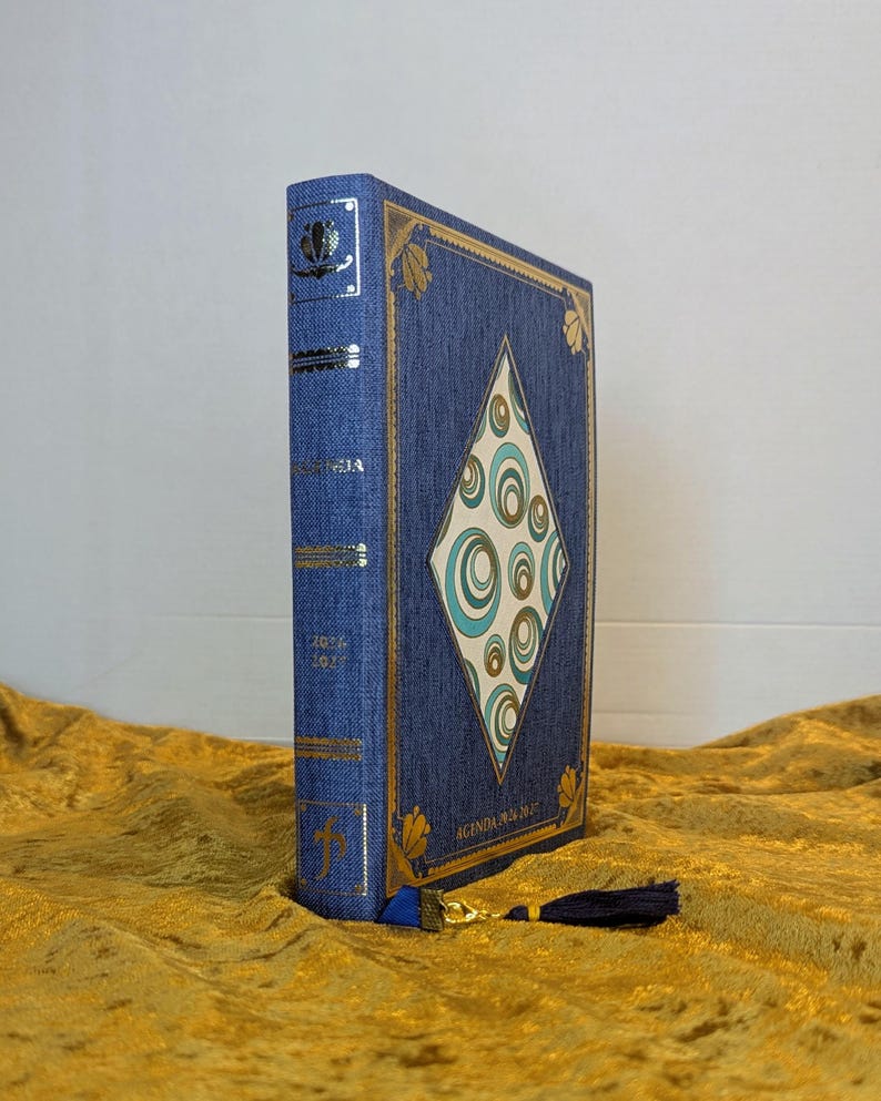 May include: A blue fabric-covered planner with a diamond-shaped centre panel featuring teal and gold concentric circles. The spine and cover are accented with gold foil detailing, including the text "AGENDA 2024-2025". A dark blue tassel is attached.