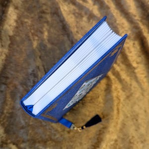 May include: A blue hardback book with a textured fabric cover, gold foil corner accents, and a white paper edge. The book features a blue ribbon bookmark attached to the spine, which is adorned with a small dark blue tassel and a gold metal charm.