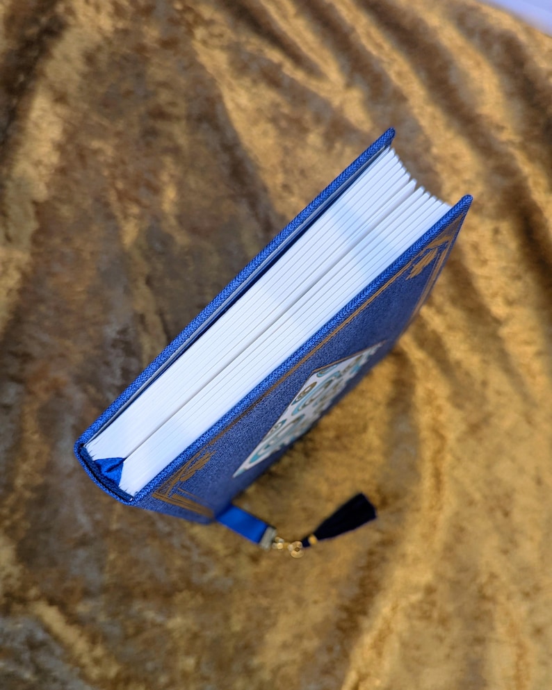 May include: A blue hardback book with a textured fabric cover, gold foil corner accents, and a white paper edge. The book features a blue ribbon bookmark attached to the spine, which is adorned with a small dark blue tassel and a gold metal charm.