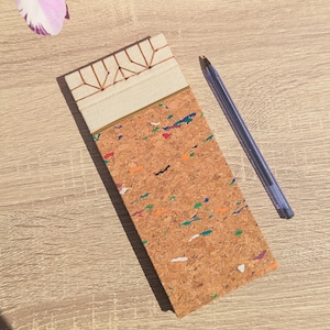 Può includere: Un taccuino rettangolare con copertina in sughero caratterizzata da macchie multicolori blu, verdi, arancioni e viola. La parte superiore presenta un dorso in tessuto color crema con cuciture geometriche color rame a vista, accompagnato da una penna blu.