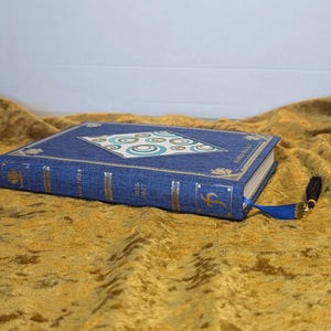 May include: A blue fabric-covered planner with gold foil accents on the spine and cover. The cover features a central diamond-shaped inlay with a teal and white circular pattern. A blue ribbon bookmark with a black tassel is attached to the spine.