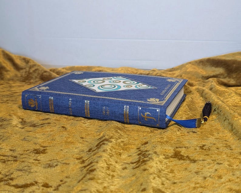 May include: A blue fabric-covered planner with gold foil accents on the spine and cover. The cover features a central diamond-shaped inlay with a teal and white circular pattern. A blue ribbon bookmark with a black tassel is attached to the spine.