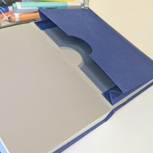 May include: A blue fabric-covered notebook featuring an interior pocket on the inside back cover. The pocket is made of dark blue cardstock with a semi-circular cutout at the top edge. The notebook pages are light grey and have a smooth, matte finish.