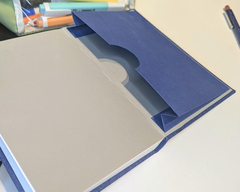 May include: A blue fabric-covered notebook featuring an interior pocket on the inside back cover. The pocket is made of dark blue cardstock with a semi-circular cutout at the top edge. The notebook pages are light grey and have a smooth, matte finish.