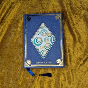 May include: A blue fabric-covered agenda for 2026-2027 featuring a central diamond-shaped inlay with a teal and gold circular pattern. The cover is decorated with gold foil borders and corner accents, and includes a blue ribbon bookmark with a tassel.