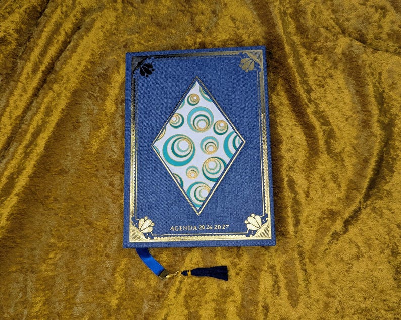 May include: A blue fabric-covered agenda for 2026-2027 featuring a central diamond-shaped inlay with a teal and gold circular pattern. The cover is decorated with gold foil borders and corner accents, and includes a blue ribbon bookmark with a tassel.