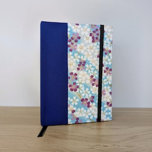 Può includere: Un taccuino rettangolare con dorso in tessuto blu scuro a tinta unita e copertina anteriore decorata con uno sfondo azzurro e un motivo ripetuto di fiori di ciliegio bianchi e viola, fissato da un elastico nero posizionato verticalmente.