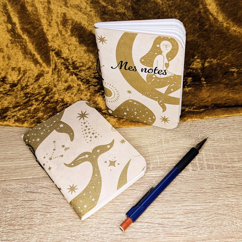 K&ouml;nnte beinhalten: Zwei Notizb&uuml;cher mit cremefarbenen Einb&auml;nden und goldenen Himmelsmustern, die eine Meerjungfrau auf dem Mond und einen Walschwanz zeigen, mit dem Text Mes notes auf einem Einband, dazu ein blauer Druckbleistift mit schwarzem Griff.