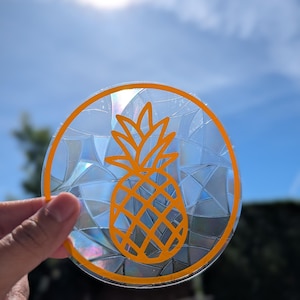 May include: A circular suncatcher featuring a vibrant orange pineapple outline centred within a circular border. The background of the design consists of a translucent, iridescent stained glass pattern that reflects light in various colours.