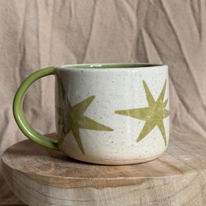May include: A ceramic mug with a speckled off-white exterior and a solid light green interior. The mug features a light green handle and a repeating star pattern in the same light green colour around the outer surface of the cylindrical body.