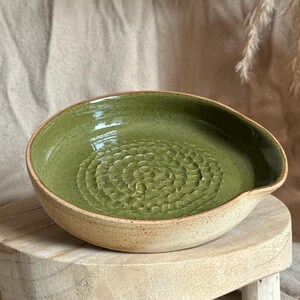 May include: A round ceramic grater plate with a speckled olive green glazed interior featuring a raised, circular, textured pattern in the centre for grating. The exterior of the plate has a natural, unglazed, light beige speckled clay finish.
