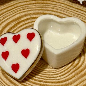 Puede incluir: Una caja de baratijas en forma de corazón con una tapa que presenta seis corazones rojos sobre un fondo blanco. La caja está abierta, revelando un interior blanco. La tapa tiene un borde dorado. La caja está sobre una superficie de madera.